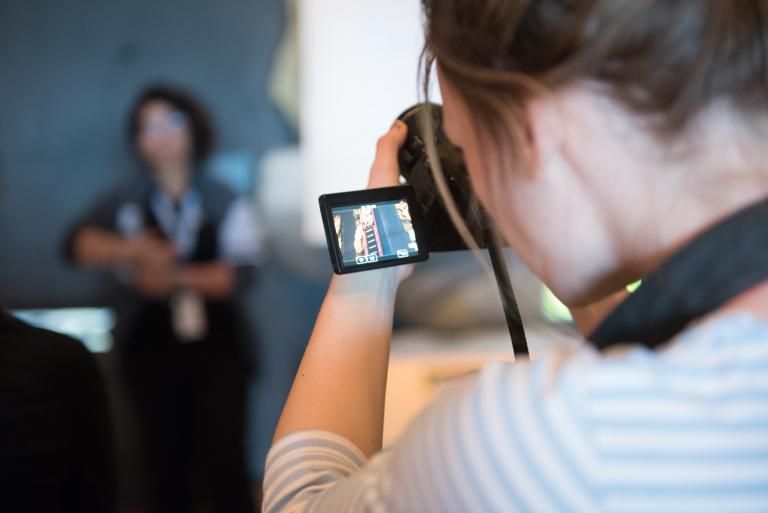 A person looks through the screen of a camera in the foreground while a person poses in the background. Partially obscured.