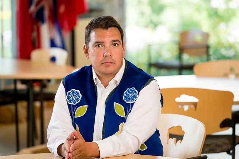 A man wearing a blue vest with flower beadwork, seated with his hands resting on a table. Partially obscured.