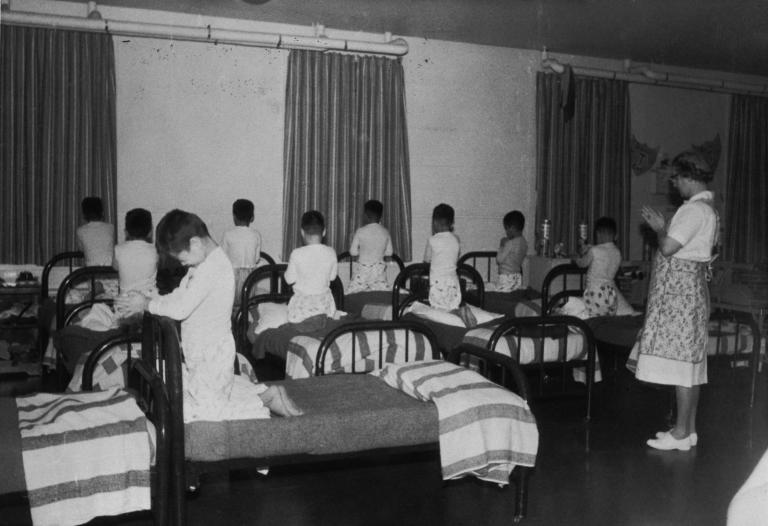 A group of boys in pyjamas kneel on single beds, heads bowed and hands clasped as if in prayer. A woman stands in the room, her hands clasped in the same way. Partially obscured.