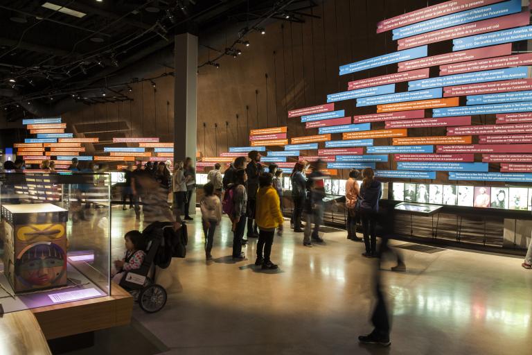 A large number of people in a gallery with a wall of colourful panels and photos showing a timeline of events. Partially obscured.