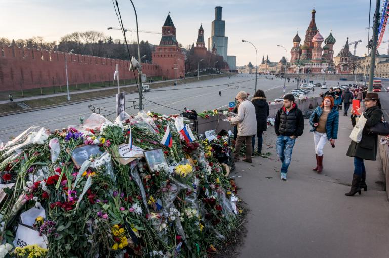 A huge pile of flowers, photographs and messages sits on a bridge beside a road. People are walking over the bridge. Some are looking at the pile of mementos. One woman is taking a photograph with her tablet. In the background is a red concrete wall (Red Square in Moscow) and a church with curvy, domed spires (St. Basil’s Cathedral).