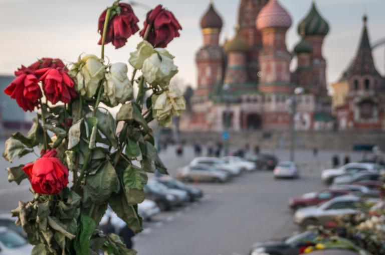 Des roses rouges et blanches fanées dans un vase. En arrière-plan, on voit la cathédrale Saint-Basile sur la place Rouge, à Moscou, avec ses multiples coupoles et dômes, qui rappellent un feu de camp montant vers le ciel. Partially obscured.