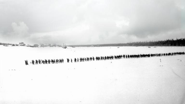 A black-and-white photo of a flat, snowy landscape with trees and small buildings in the distance. A long, straight line of more than 100 people are walking through snow. Partially obscured.