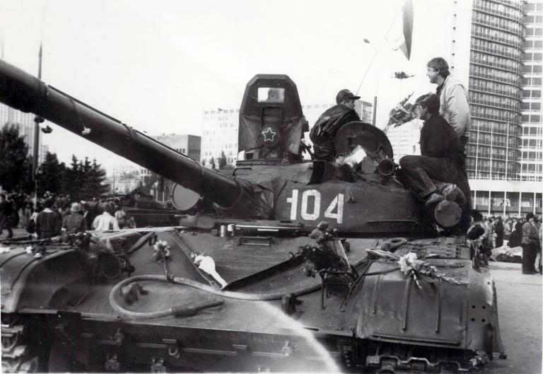 Two men stand on a parked military tank, talking. There are bouquets of flowers strewn across the tank. Behind are people milling in front of a tall building. The photo is black and white.