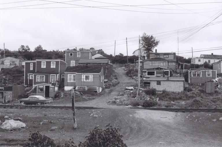 A black and white image of about a dozen houses on a hill. In the middle of the image, a dirt road travels up the hill between the houses.