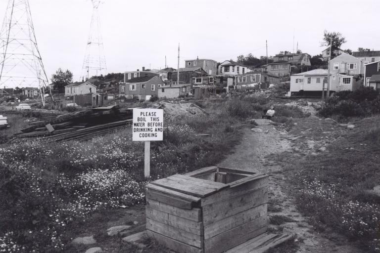 A black and white image of about 15 houses on a hill. In the foreground is a well with a sign beside it which reads: “Please boil this water before drinking and cooking.