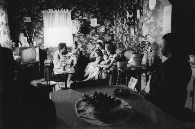 A black and white image of women and young children sitting on couches and chairs in a small living room.