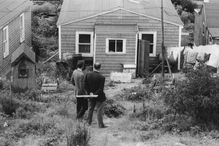 A black-and-white image of two men in suits standing in front of a wooden house with their backs to the viewer. 