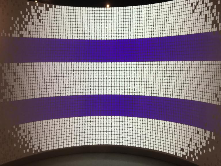 An image being projected onto a curved wall that is about nine feet tall. It is made up of many small squares and has the appearance of Indigenous bead work or a mosaic. The image is mainly made up of white squares, but it also features two thick blue horizontal lines that run parallel to each other. Partially obscured.