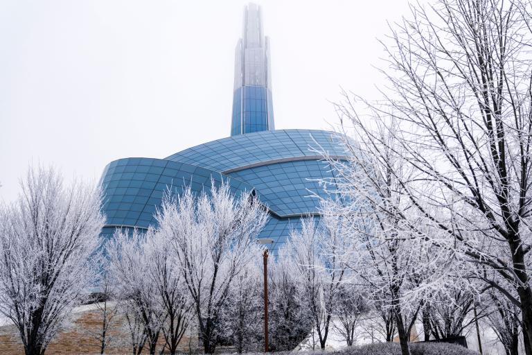 An unusual building surrounded by a glass "cloud" and topped by a tower. It is surrounded by snow and bare trees. Partially obscured.