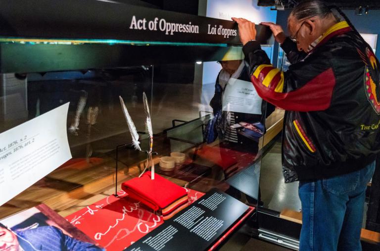 A man with long, braided hair bows his head in front of a glass-walled display where two feathers are lodged. Partially obscured.