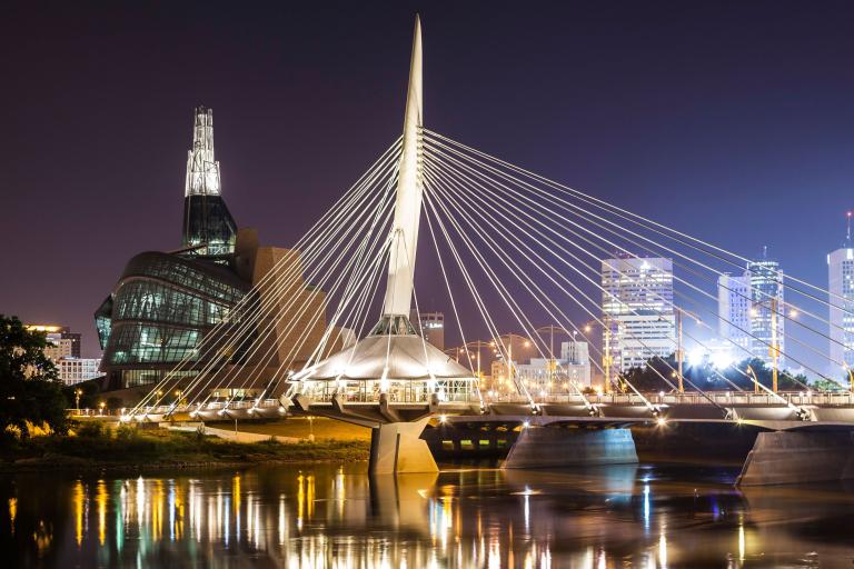A nighttime shot of an unusual building with a glowing tower, standing beside an illuminated modern bridge, which is reflected in the river water below. Partially obscured.