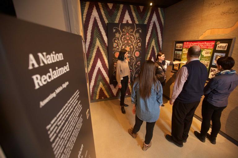 A guide speaks to a small group of people in a museum exhibit space. The space includes a wall featuring a Métis artwork and a text panel that reads “A Nation Reclaimed”. Partially obscured.