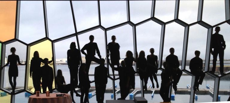 Eighteen people in a row, some seated, some standing, in front of a wall of odd-shaped windows. Partially obscured.