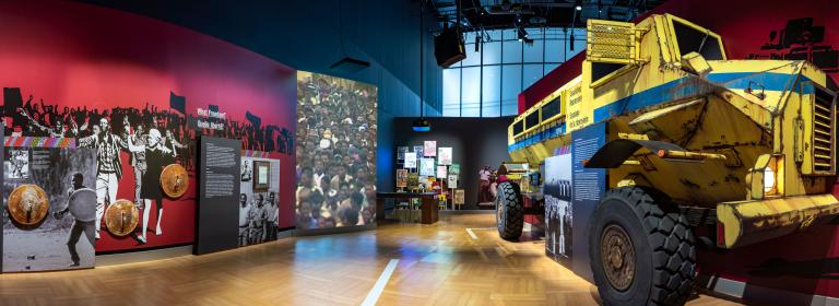 A museum gallery with large, yellow armoured truck emerging from one wall and rusty garbage-can lids hanging on the opposite wall. Partially obscured.