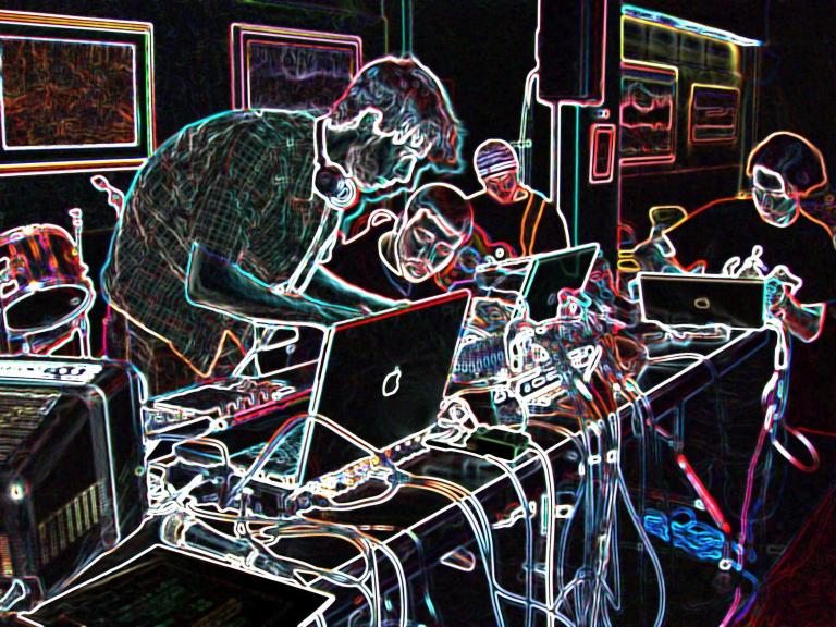 Four people around a table covered in computers and electronic music equipment. The photo has been altered so that the background is black and the elements of the photo are outlined in multi-coloured neon. Partially obscured.