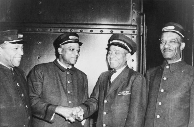 A black and white photo of four men in train porter uniforms. All of the men are smiling, and the two men in the middle appear to be shaking hands. Partially obscured.