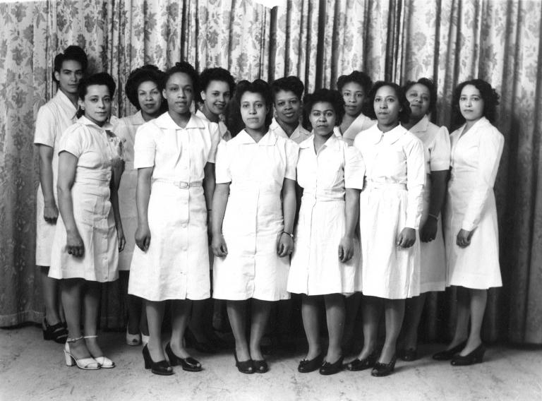 A black and white image of Viola Desmond with 11 other women. All of the women are wearing plain white dresses and are standing in two rows, posing for the camera.