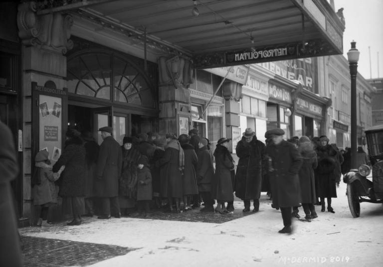 A black and white image of a crowd in front of a theatre entrance. The theatre is named “Metropolitan”. Everyone is wearing long old-fashioned coats, and some are wearing hats. An antique car is parked on the street.