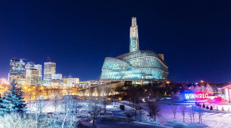 A glass and steel building on a snowy evening, positioned behind an illuminated sign that says Winnipeg. Partially obscured.
