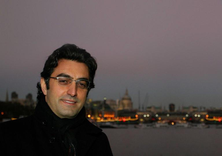 Close-up of a smiling man at dusk. Behind him is a body of water and on the other side there are boats, lights and buildings. Partially obscured.