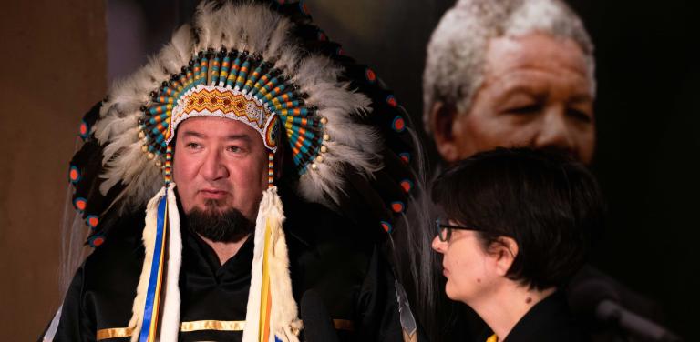 A man wearing a feather headdress stands beside a woman, who is looking at him. There is a large photograph of a man with grey hair (Nelson Mandela) in the background. Partially obscured.