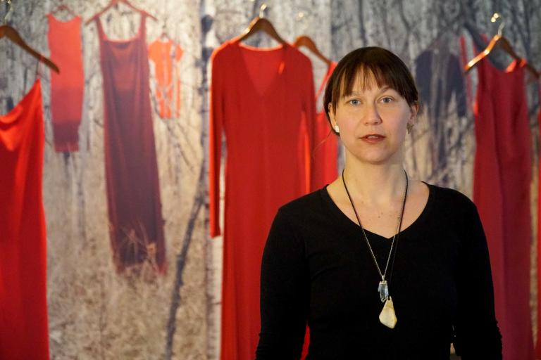 Jaime Black stands in front of red dresses suspended in air on hangers in front of a backdrop. The backdrop features an image of a birch wood forest with more red dresses hanging in it. Partially obscured.