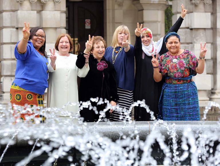 Six women are standing in front of what appears to be the entrance of a large stone building. They are all smiling and holding up one or two hands making the peace sign.