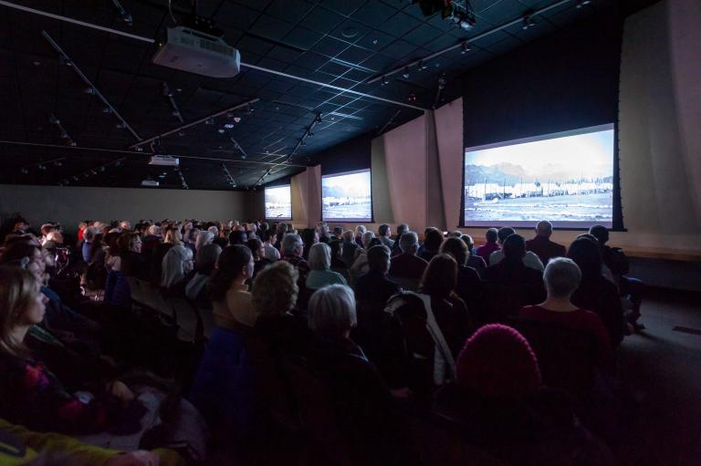 A seated audience viewing three large screens in a dimly lit room. Partially obscured.