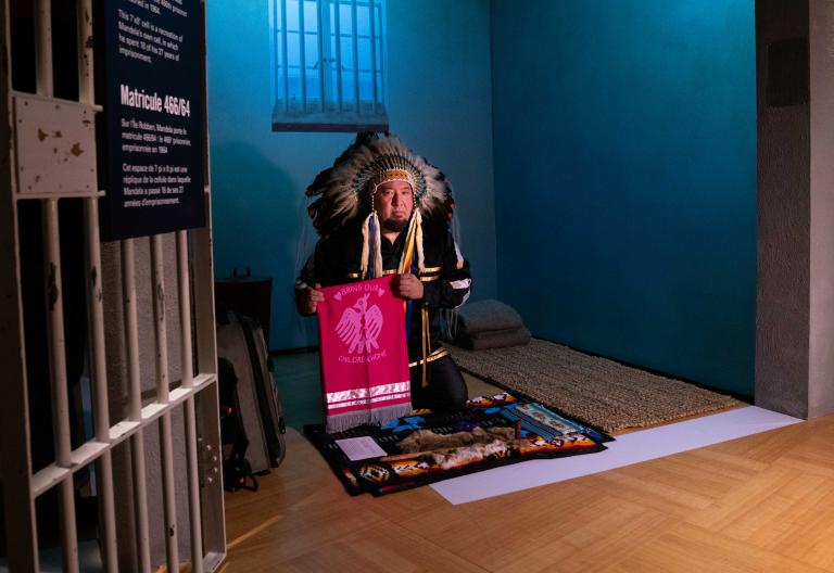 An Indigenous man wearing ceremonial regalia kneels inside a replica of a prison cell. He is presenting a textile banner with a graphic image of a bird and the words “bring our children home.” Partially obscured.