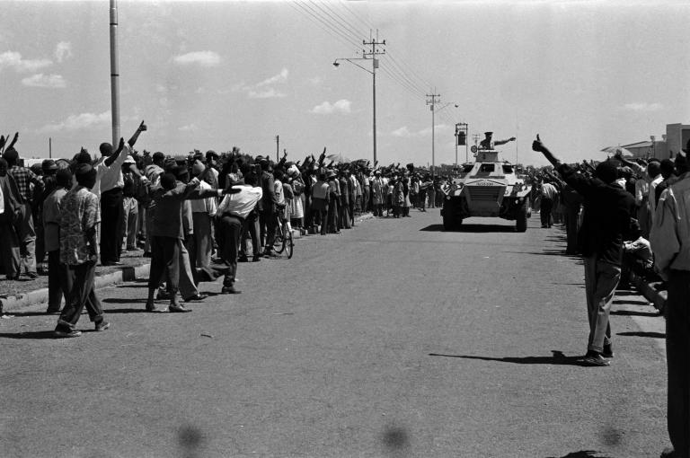 A large armoured vehicle drives down a road. Crowds on either side of the vehicle hold out their fists in a salute with their thumbs raised.