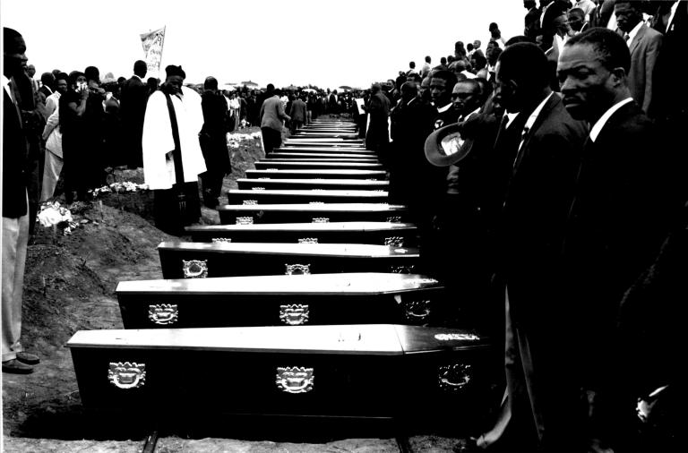 People stand in front of a row of coffins. Partially obscured.