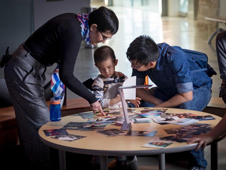 A woman, a man and a young boy are gathered around a circular table. Several photographs are scattered on the table, and the woman is pointing one out to the boy. Partially obscured.