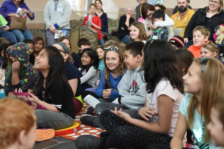 Smiling children sitting cross-legged on the floor. One girl in the centre is looking into the camera. Partially obscured.