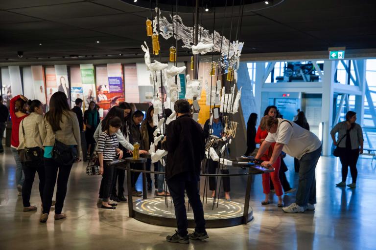 A large number of people in a gallery examining a display comprised of several seemingly ordinary objects Partially obscured.