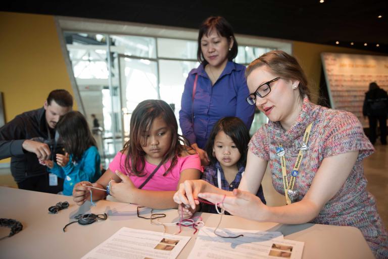 A woman strings yarn around her fingers as she demonstrates and explains what she is doing to two girls. Partially obscured.