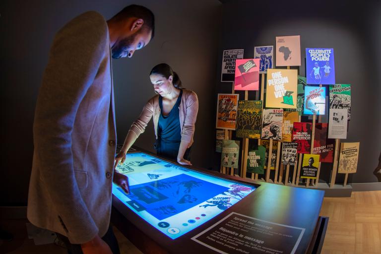 A man and woman look down at an illuminated table. In the background, there is a grouping of colourful posters on the wall. Partially obscured.