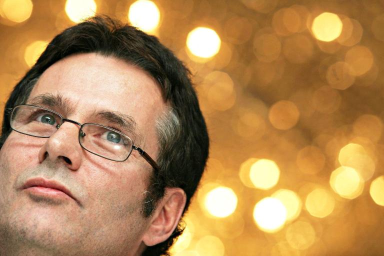 Close-up of a man’s face looking skyward in a thoughtful pose. He is wearing glasses and there are blurry spots of light in the background. Partially obscured.