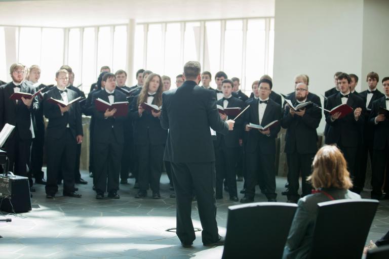 A choir of men wearing black outfits hold books and sing in front of a conductor. Partially obscured.