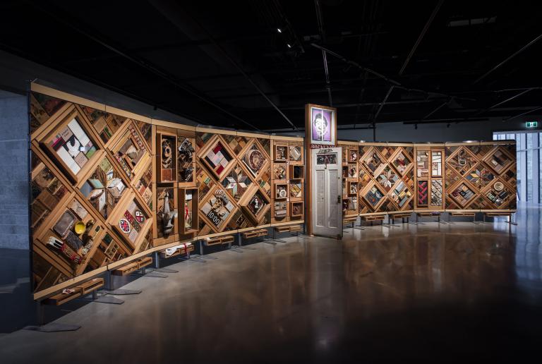 A large horizontal art installation with a wooden frame made up of multiple diamonds and rectangles to which many objects are mounted. There is a half-opened door in the middle of the work.