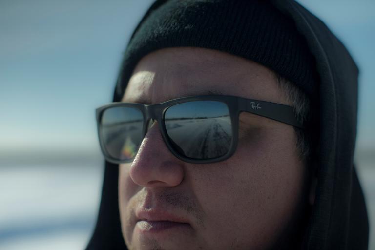A close-up of a man wearing sunglasses and a hat, looking at something off camera. A road is seen reflected in his sunglasses. Partially obscured.