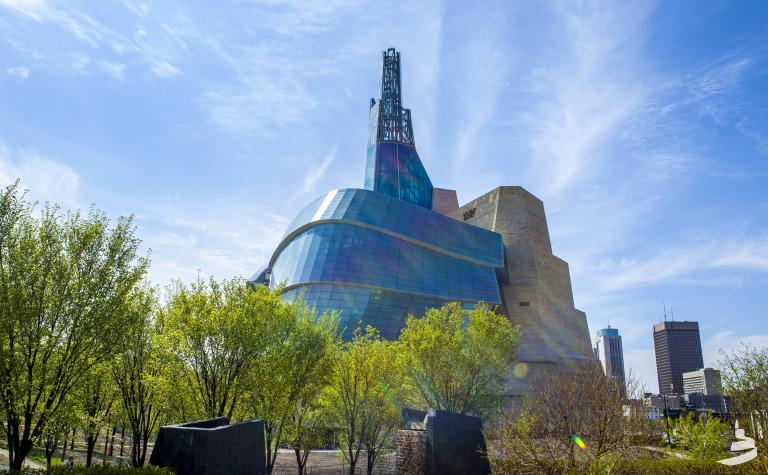 An unusual glass, steel and concrete building stands behind light green tree branches and green grass, against a bright blue sky. Partially obscured.