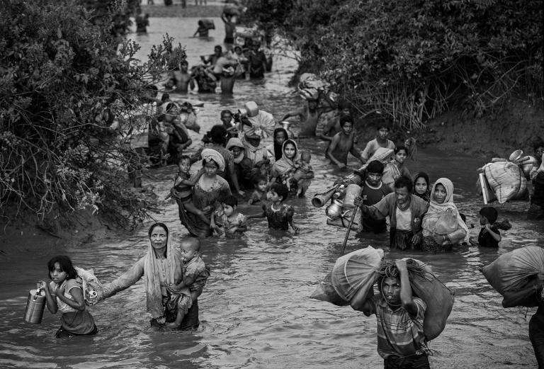 Rohingya women, children and men wade through waist-high muddy river water. Some carry young children, while others carry bags of possessions, including household items. Partially obscured.