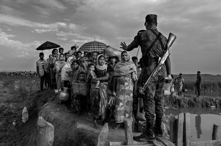 A soldier with a rifle on his back blocks a group of Rohingya before they cross a makeshift bridge. His left hand is raised, gesturing for them to stop.