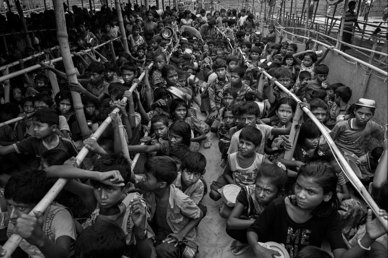 Hundreds of Rohingya children, most of them crouched or sitting down, wait in long lines for food. The line-ups are separated by long rows of bamboo poles.