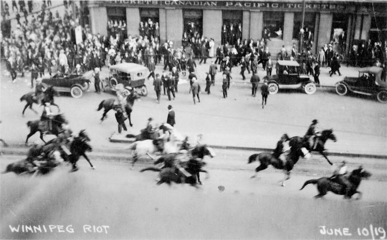 A group of men on horseback ride down a city street as a crowd looks on from the sidewalk.