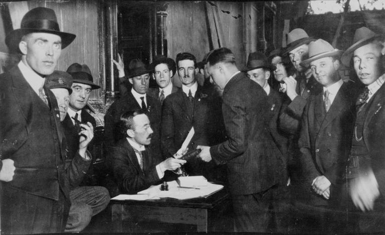 A group of men crowd around a man at a desk.