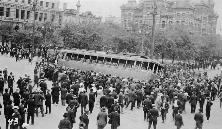 A large crowd of people trying to tip over a streetcar.