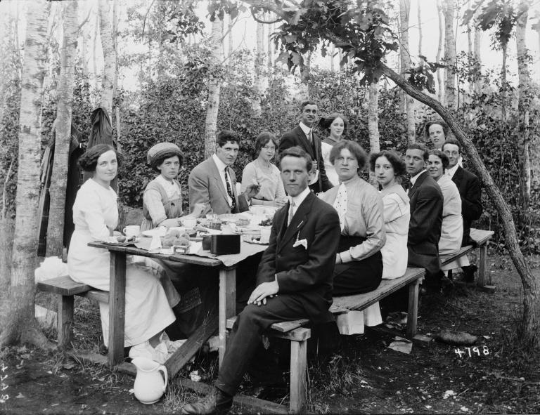 A group of well-dressed men and women sitting at a picnic table with trees in the background.
