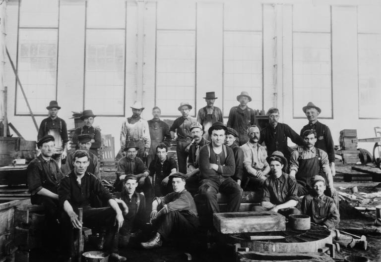 A group of men in working clothes sitting and standing in a factory.